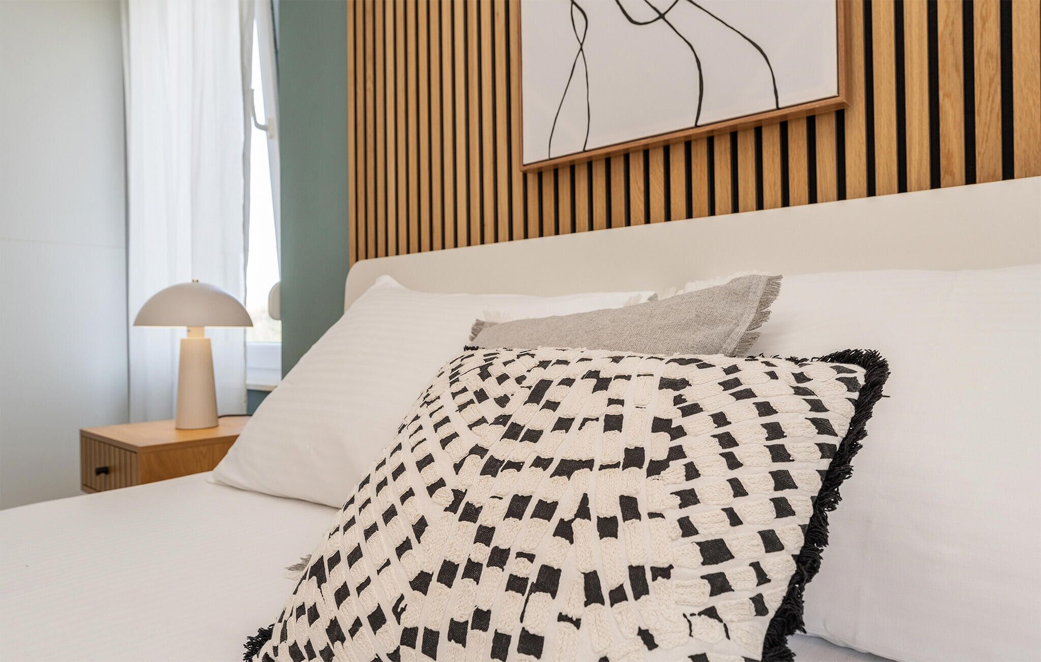Un lit bien fait, agrémenté d’un coussin décoratif à motifs et d’un coussin uni, une lampe de chevet et un mur avec des lattes de bois verticales.