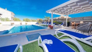 A pool area with blue lounge chairs, a covered seating area, and a view of surrounding buildings. - Stunning apartment in Bibinje (Bibinje)