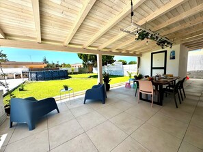 A covered patio area with a tiled floor, blue chairs, a wooden table, and a view of a well-maintained lawn and white walls. - Mags House a 300 mt dal Mare (Santa Maria del Focallo)