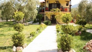 Um edifício de dois andares com varanda, rodeado por um jardim bem cuidado com árvores e arbustos.