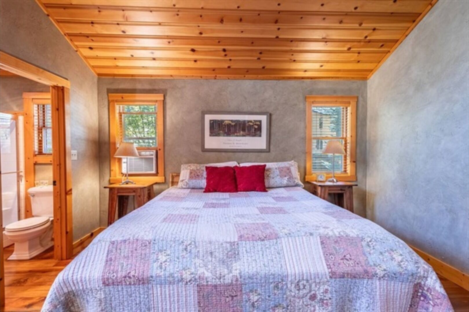 A bedroom with a wooden ceiling, a bed with a quilt, two bedside tables, and a window.