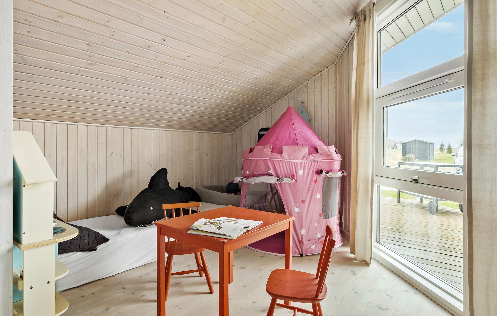 Ein gemütliches Zimmer mit Holzbalkendecke, einem rosa Spielhäuschen, einem Tisch mit Stühlen und einem Fenster mit Blick ins Freie.