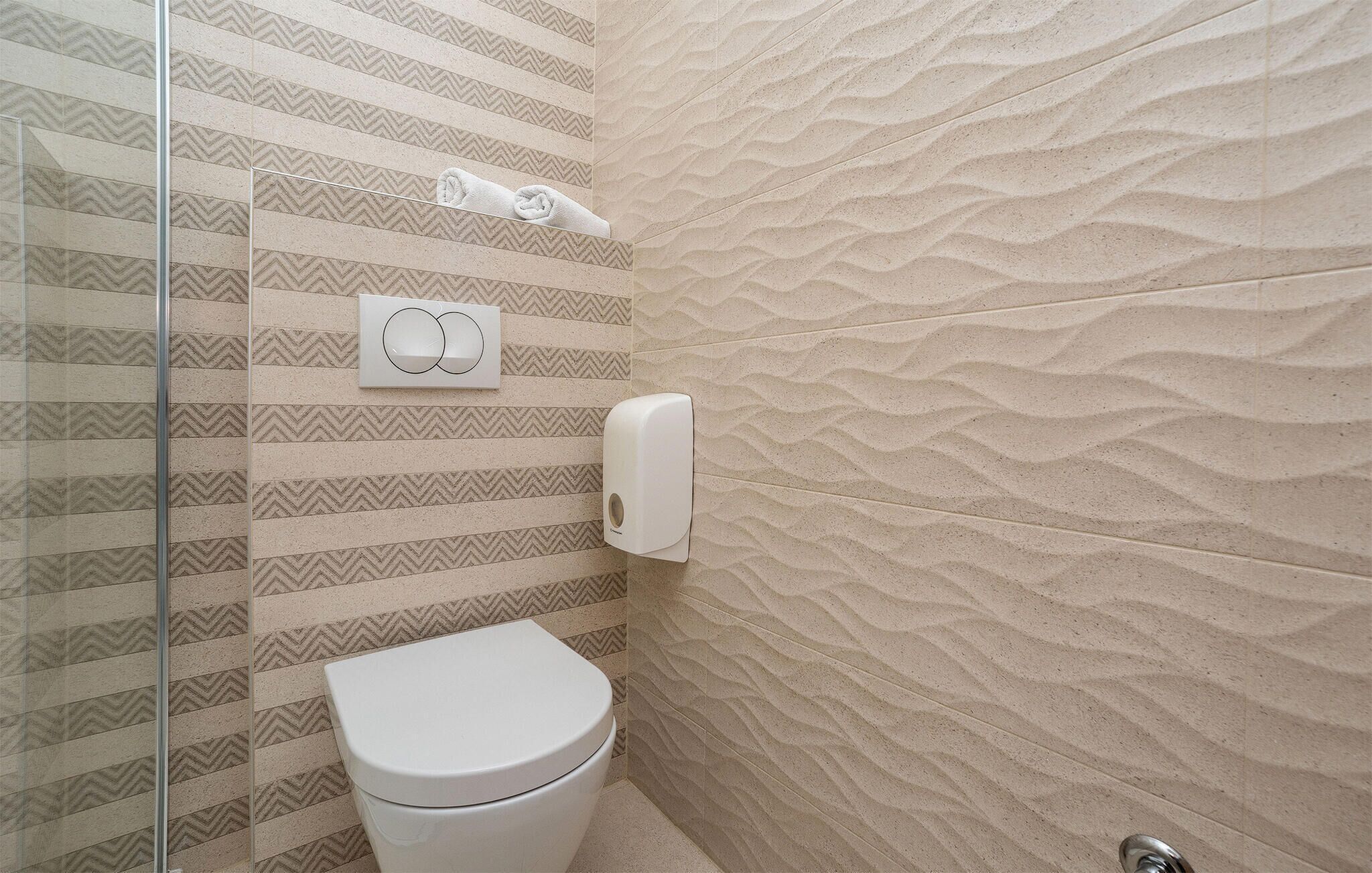 A bathroom with a striped tile wall, a toilet, a towel dispenser, and a glass shower enclosure.
