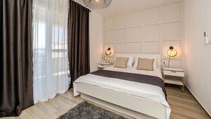 Ein Schlafzimmer mit einem großen Bett, zwei Nachttischen, einem modernen Kronleuchter und Blick durch transparente Vorhänge.