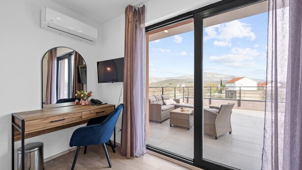 Ein modernes Hotelzimmer mit einem Holztisch, einem blauen Sessel, einem Flachbildfernseher, einer großen Schiebeglasdoor zum Balkon mit Sitzgelegenheit und Blick ins Grüne.