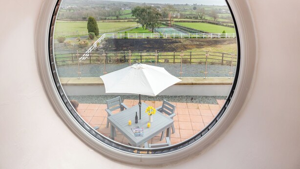 Une fenêtre ronde donnant sur un champ verdoyant, une clôture blanche et une table avec des chaises et un parasol.
