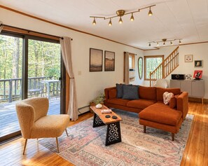Un salon chaleureux avec un canapé, un fauteuil et une table basse, un escalier et une vue sur les arbres à travers de grandes fenêtres.