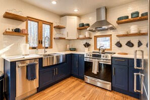 Une cuisine moderne avec des meubles bleu foncé, des appareils en acier inoxydable, des étagères en bois et une fenêtre donnant sur les arbres.