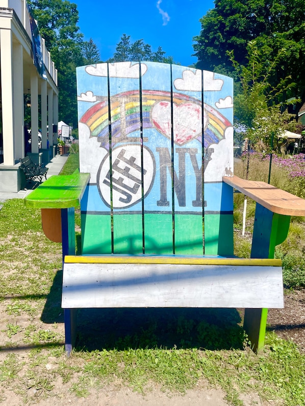 Una silla Adirondack colorida con un diseño pintado, que incluye un arcoíris y nubes, ubicada en un área con césped.