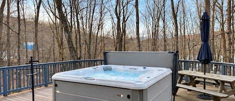 A hot tub on a wooden deck with a picnic table and umbrella, surrounded by leafless trees.
