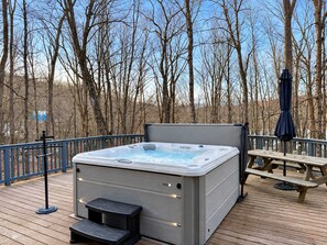 Un bain à remous sur une terrasse en bois, avec une table de pique-nique et un parasol, le tout entouré d’arbres sans feuilles.