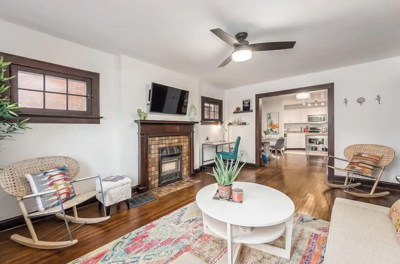 A spacious living room with a fireplace, a ceiling fan, and a view into the kitchen.