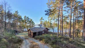 A rustic cabin with a gravel path leading to it, surrounded by tall pine trees and a clear view of a lake in the background. - 2 bedroom cozy home in Jørpeland (Jørpeland)