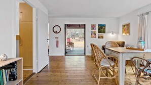 Een kamer met een houten vloer, een eettafel met stoelen, een boekenplank en een deur die leidt naar een buitenruimte met uitzicht op bomen en een rode constructie.