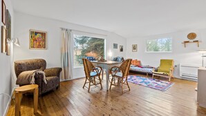 Een gezellige woonkamer met houten vloer, een eettafel met stoelen, een bank en een klein bijzettafeltje. Twee ramen zorgen voor daglicht en uitzicht op bomen.