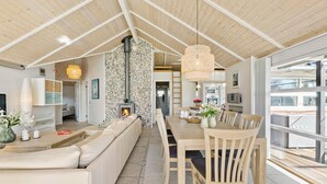 A modern living room with a stone fireplace, wooden ceiling, and a dining area with a large window. - Stunning home in Gilleleje with sauna (Gilleleje)