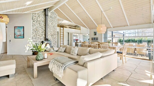 A spacious living room with a large sofa, a coffee table, and a fireplace. There is a dining area with wooden chairs and a table. The room has a high ceiling with wooden beams and large windows. - Stunning home in Gilleleje with sauna (Gilleleje)