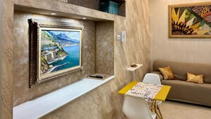 Una habitación con un cuadro de un pueblo costero, una mesita con superficie amarilla, una silla blanca y un sofá con cojines dorados.