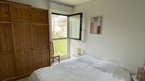 A bedroom with a large bed, a wooden wardrobe, a small table, and a window with a view of greenery.
