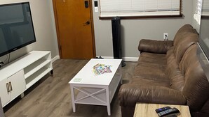 Ein Wohnzimmer mit Fernseher, einem weißen Couchtisch, einem braunen Ledersofa und einem kleinen Holztisch mit Fernbedienung.