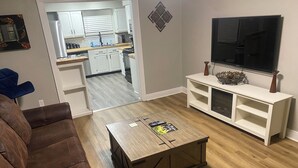 Ein Wohnzimmer mit braunem Ledersofa, einem Holztisch mit Fernbedienungen, einer weißen Unterhaltungsanlage mit Fernseher und einer durchgehenden Tür sichtbaren Küche.