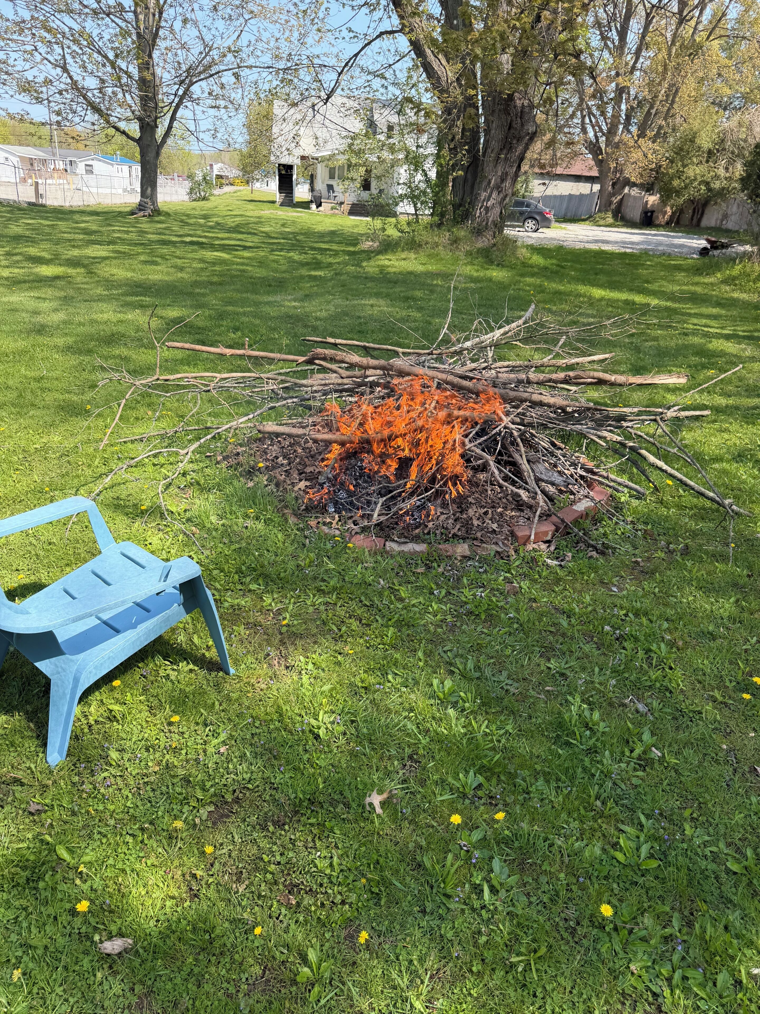 A backyard with a fire pit surrounded by grass, a blue plastic chair, and trees.