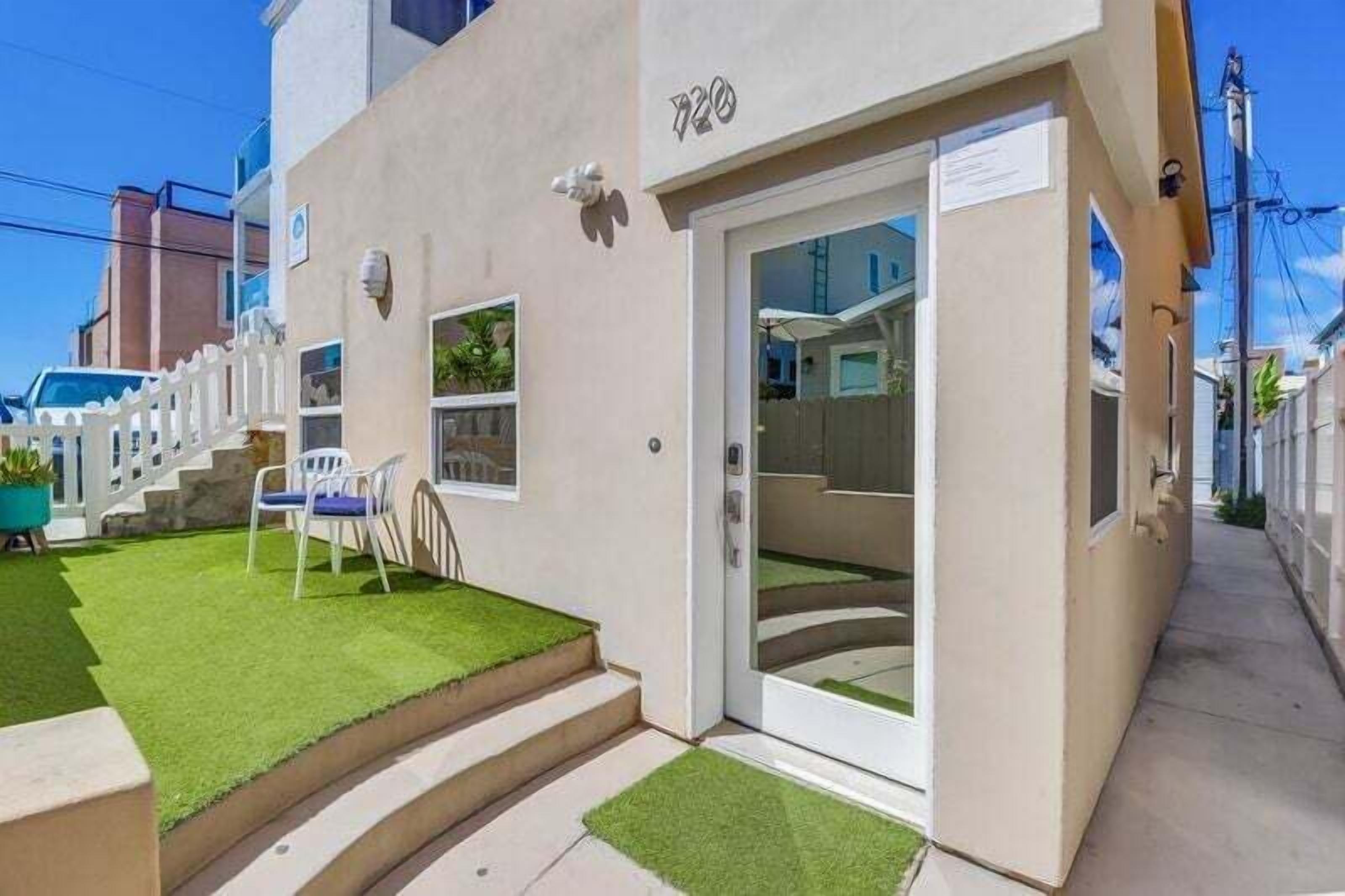 Un bâtiment moderne à la façade blanche, doté d’une porte avec des panneaux de verre et d’un petit espace extérieur aménagé avec une chaise et une table.