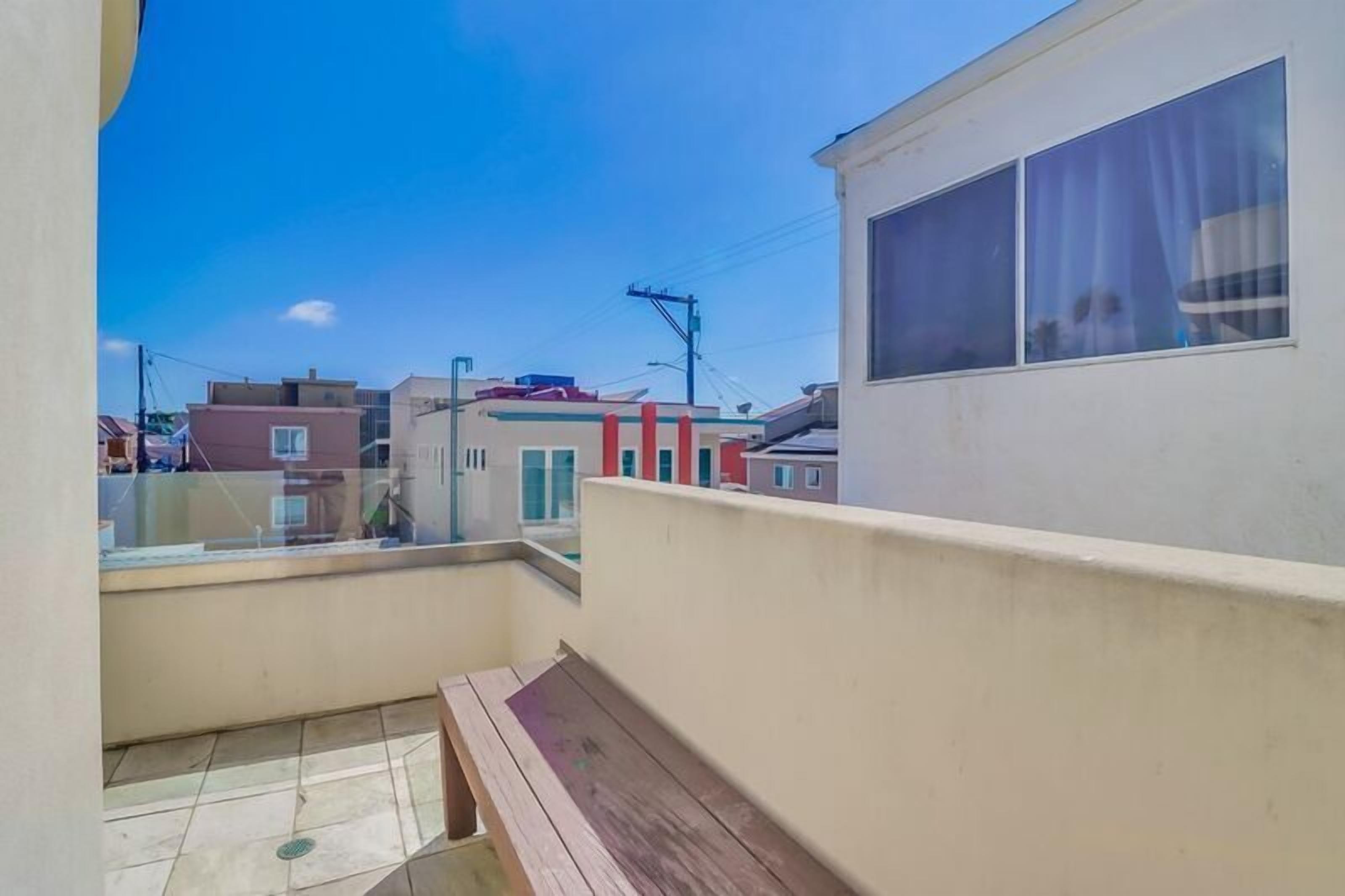 A balcony with a wooden bench, clear sky, and neighboring buildings.