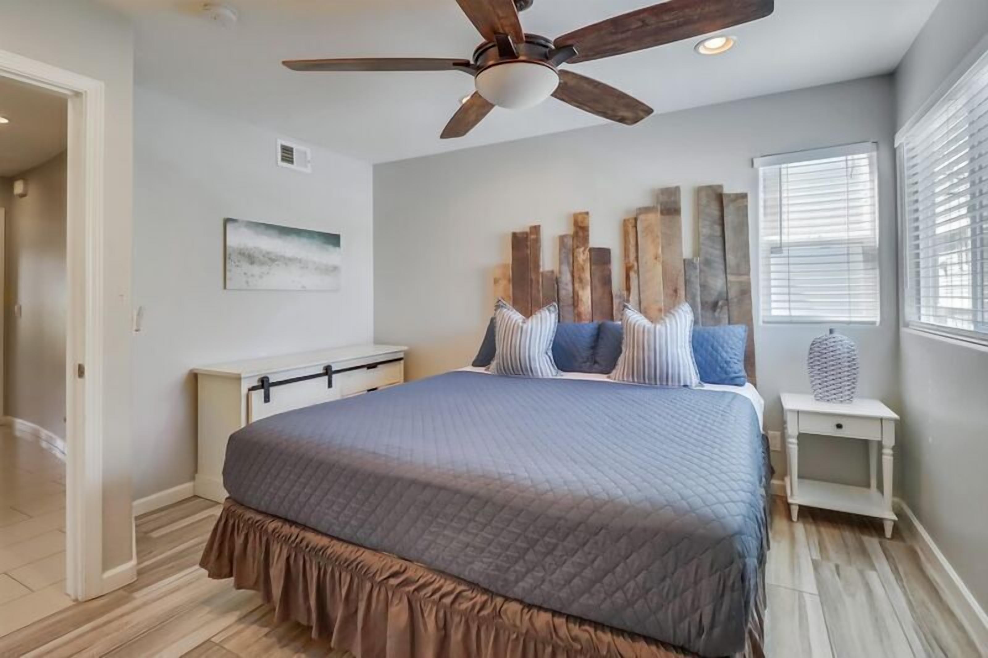 A bedroom with a large bed, a ceiling fan, a nightstand, and a window with blinds.