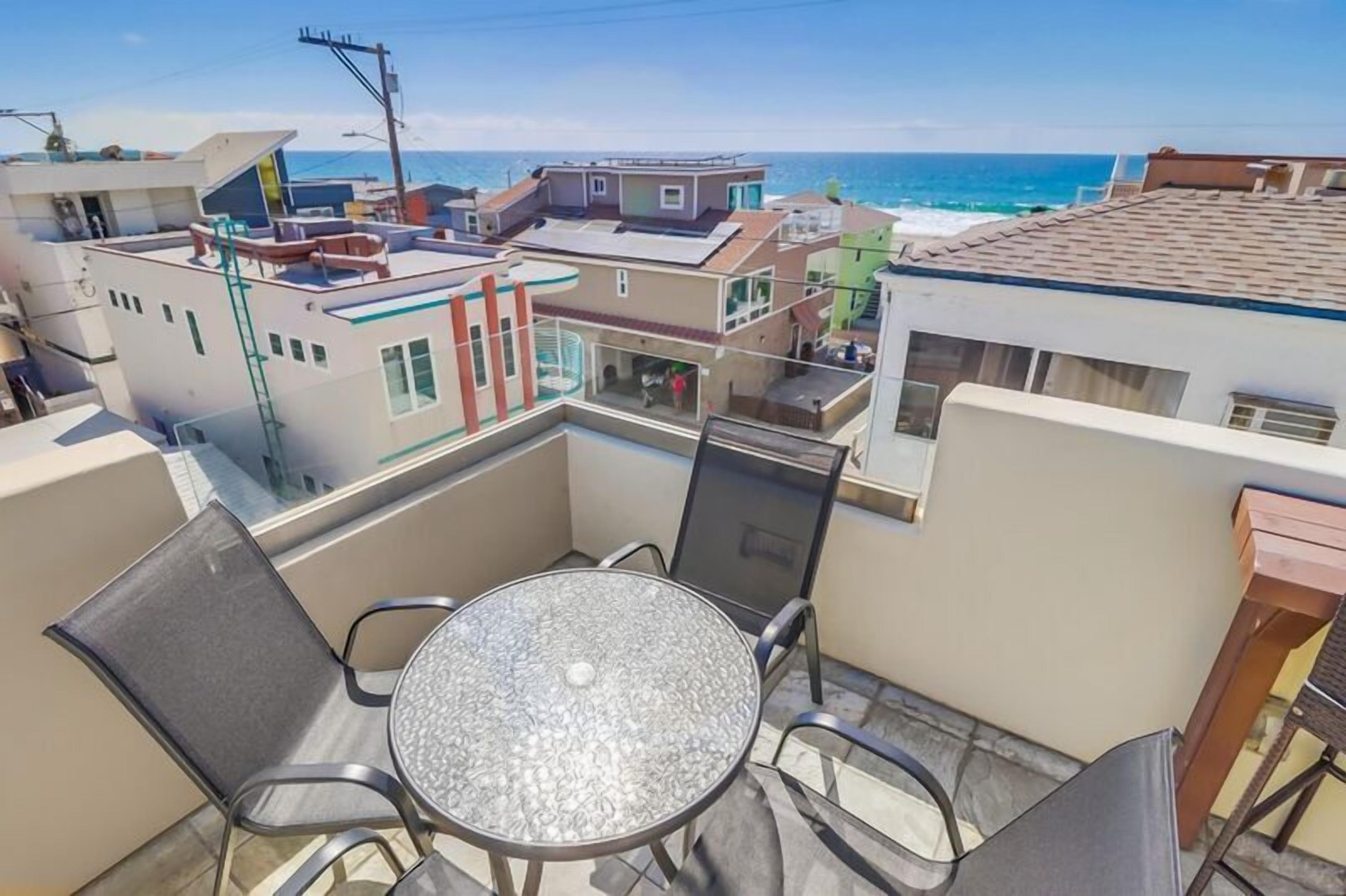 A balcony with a glass table and chairs, overlooking a coastal area with buildings and the sea.