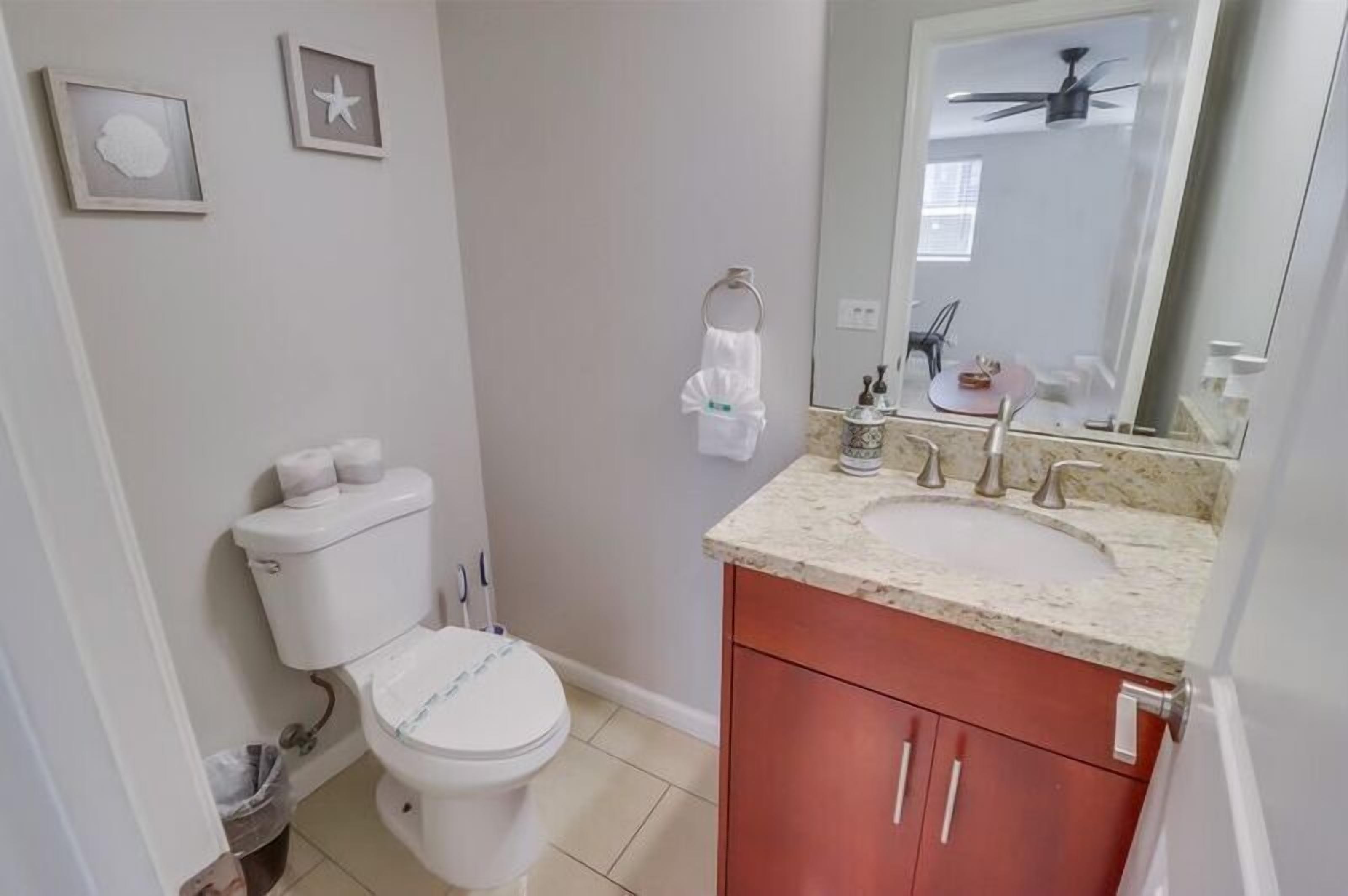 A bathroom with a toilet, sink, and mirror. There are framed pictures on the wall and a ceiling fan.