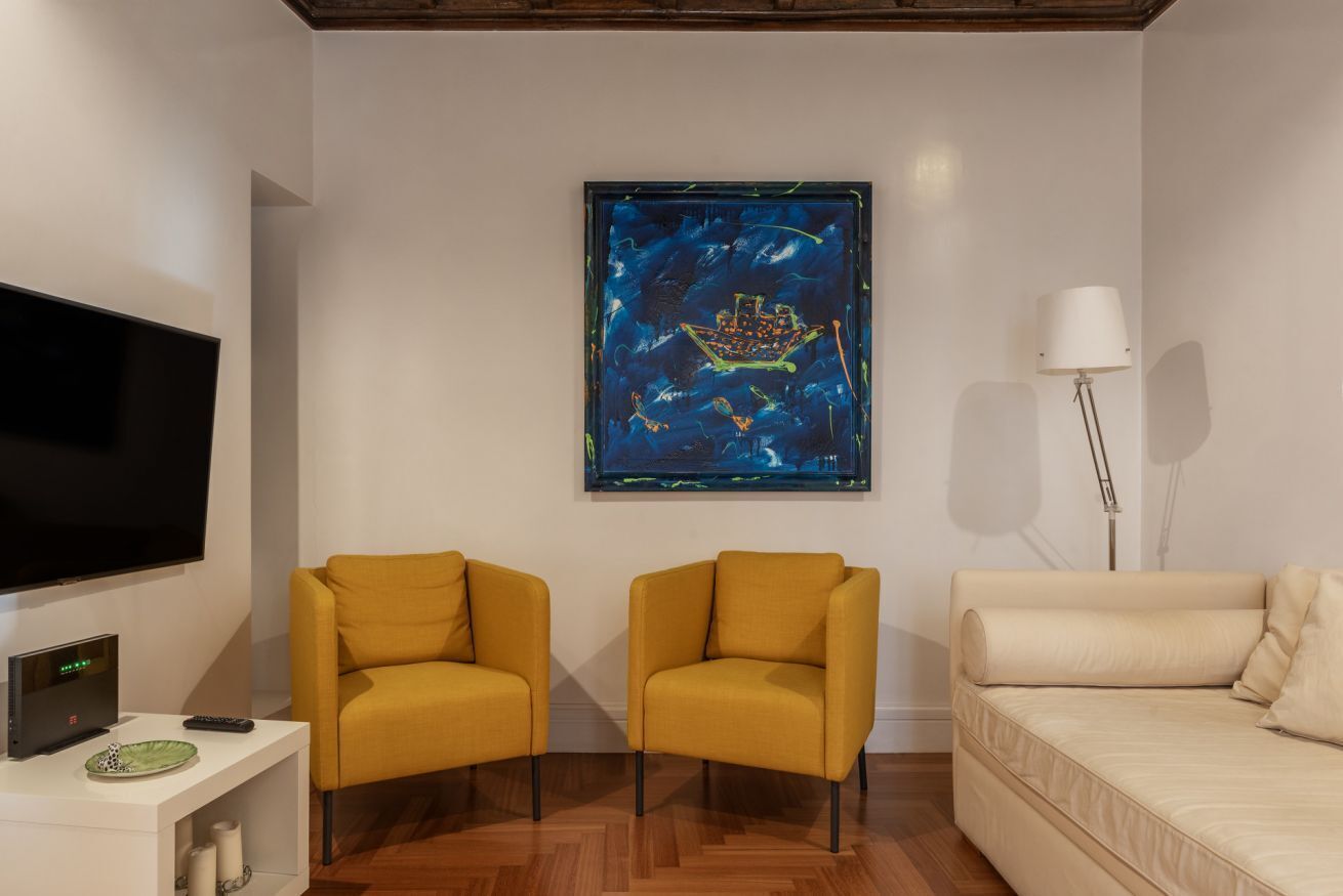 A living room with a flat-screen TV, two yellow armchairs, a beige sofa, a framed artwork on the wall, a floor lamp, and a white side table with a remote control and a plate.