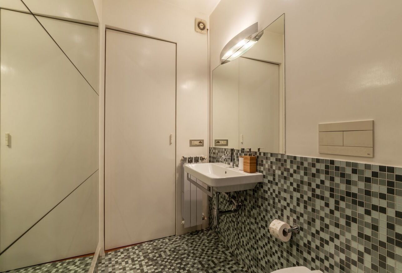A bathroom with a tiled floor, a sink, a toilet, a mirror, and a wall-mounted light fixture.