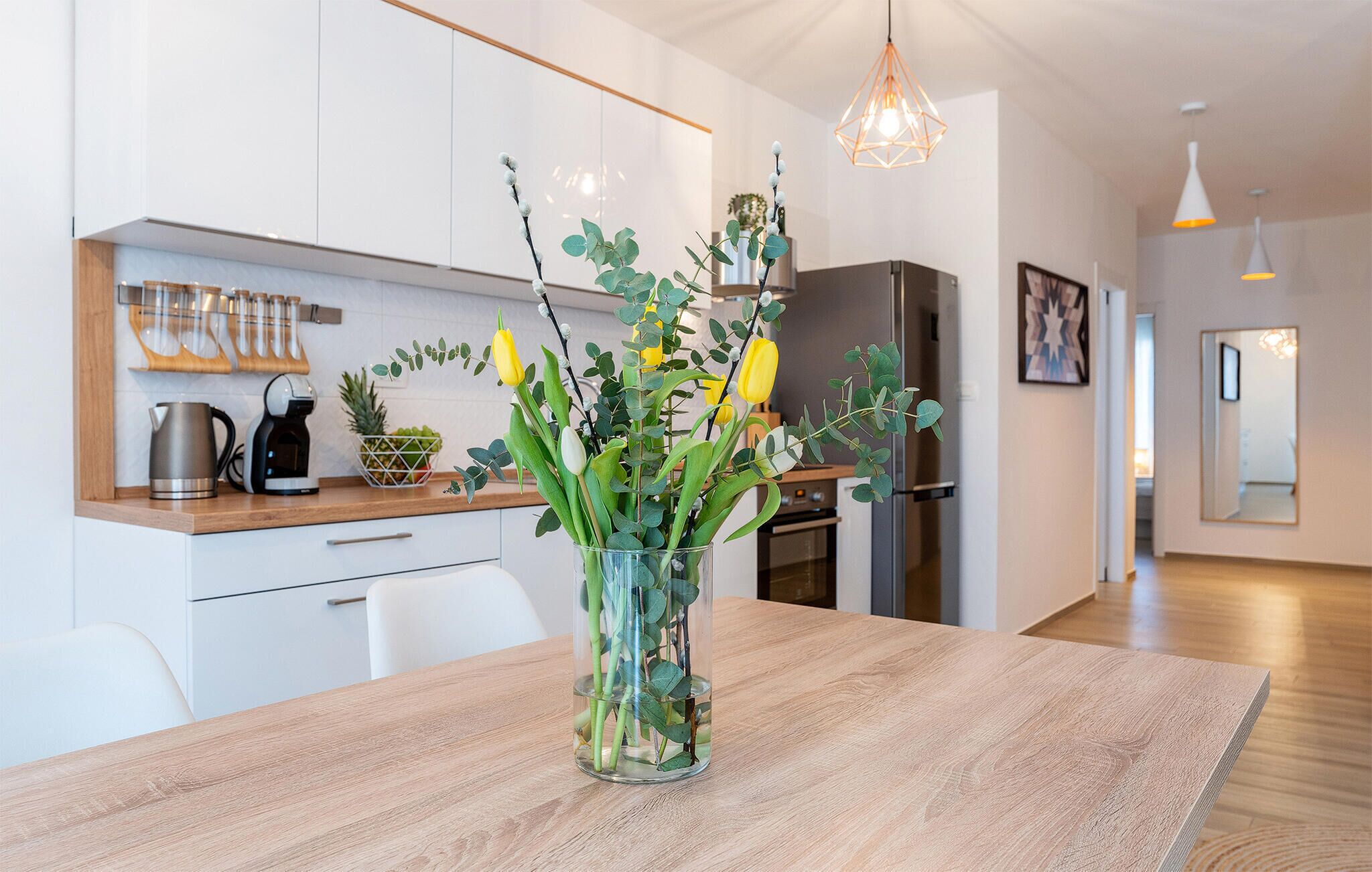 Una cocina moderna con armarios blancos, encimera de madera, cafetera y un florero con flores sobre la mesa.