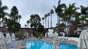 Un espace piscine agrémenté de chaises longues et de parasols, entouré de palmiers et bordé d’un bâtiment dont le toit est recouvert de tuiles rouges.