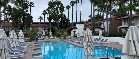 A pool area with lounge chairs and umbrellas, surrounded by palm trees and a building with a red-tiled roof.