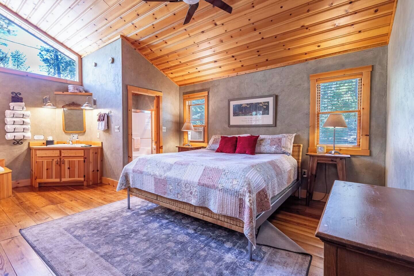 Ein Schlafzimmer mit Holzbalkendecke, einem Bett mit gemustertem Überwurf, einem Nachttisch, einem Beistelltisch mit Lampe und einem Fenster mit Jalousien.