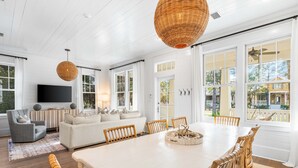 A spacious living room with a large white dining table, wicker chairs, and a grey armchair. - Vibrant WaterColor Haven: Electric Cart, Closet to Camp WaterColor, & Green Space Galore (Santa Rosa Beach)