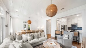 A modern living room with a sofa, armchair, and coffee table, connected to a kitchen with white cabinets and a dining area. - Vibrant WaterColor Haven: Electric Cart, Closet to Camp WaterColor, & Green Space Galore (Santa Rosa Beach)