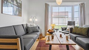 A modern living room with a grey sofa, a wooden coffee table, and a view of a house through large windows.