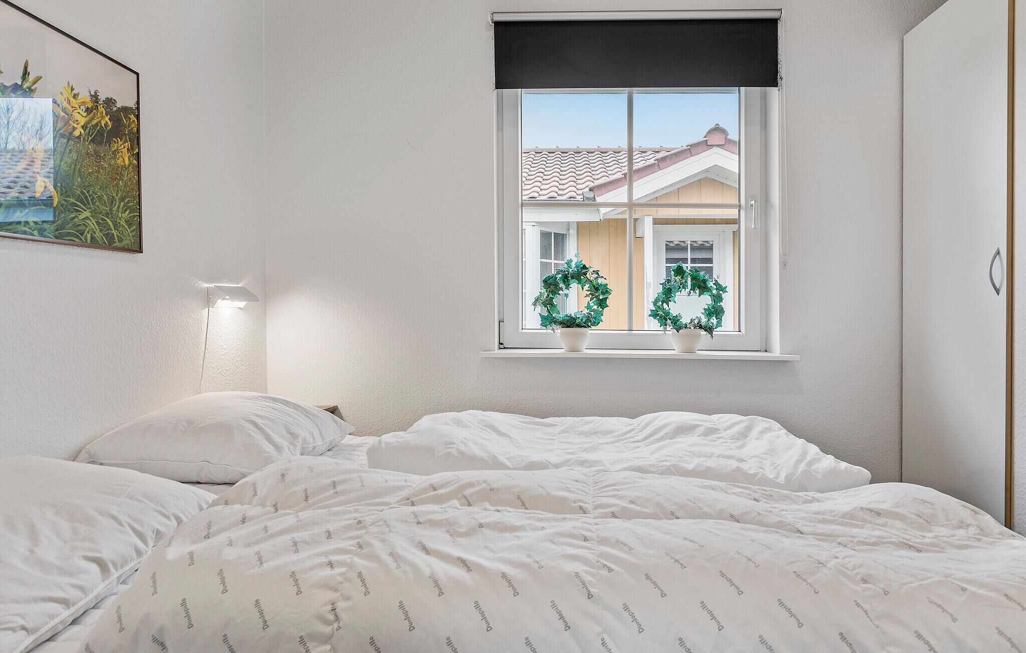 Ein Schlafzimmer mit einem Bett, einem Fenster mit Blick auf ein Haus und einer Wandleuchte.