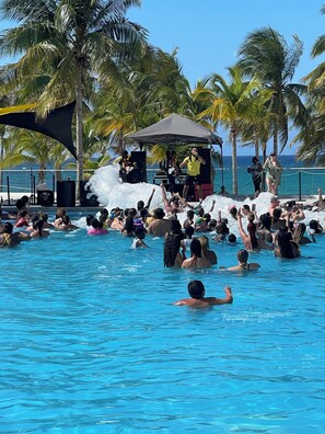 A crowded pool area with people enjoying the water, surrounded by palm trees and a stage with performers.