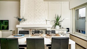 Un coin repas agrémenté d’une cheminée en briques blanches, d’une plante en pot et d’une table dressée pour quatre personnes.