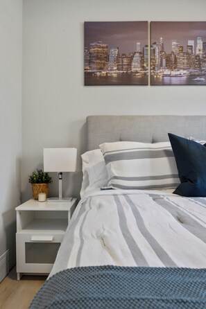 A neatly arranged bedroom with a bed, pillows, a bedside table with a lamp, and a cityscape painting on the wall.