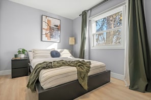 Un dormitorio con cama, mesita de noche, lámpara, ventana con cortinas y una obra de arte en la pared.