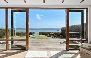 A modern room with large glass doors leading to a wooden deck overlooking a body of water. - 2 bedroom beach front home in Rønde (Rønde)