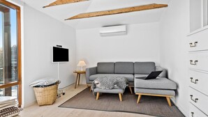Ett modernt vardagsrum med en grå soffa, ett litet sidobord med en lampa, en tv som är monterad på väggen och ett stort fönster med utsikt över staden.