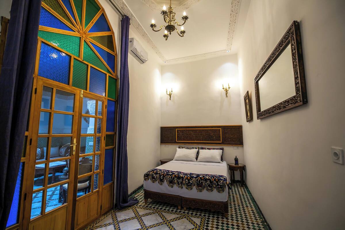 A bedroom with a large bed, a wooden door with glass panels, a chandelier, and a wall-mounted mirror.