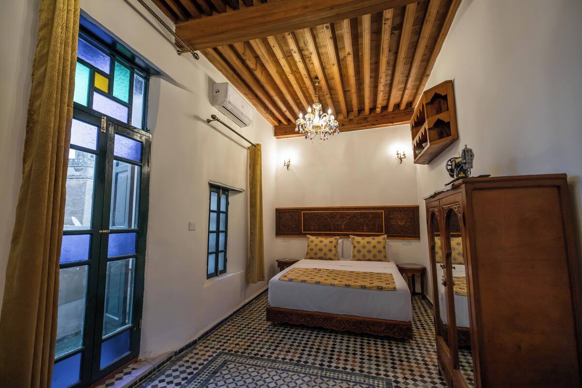 A bedroom with a large bed, wooden headboard, a chandelier, a wardrobe, and a patterned floor.