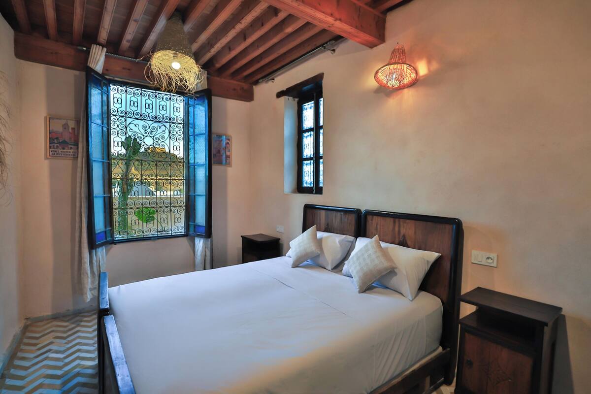 A bedroom with a wooden bed frame, a large window with decorative iron bars, and a small bedside table.